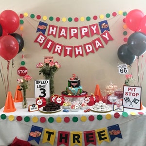 May include: A Cars-themed birthday party table setting with red and black balloons, a cake with a race car on top, and a banner that says "Happy Birthday". There are also various decorations, including traffic signs, cones, and a banner that says "Three".