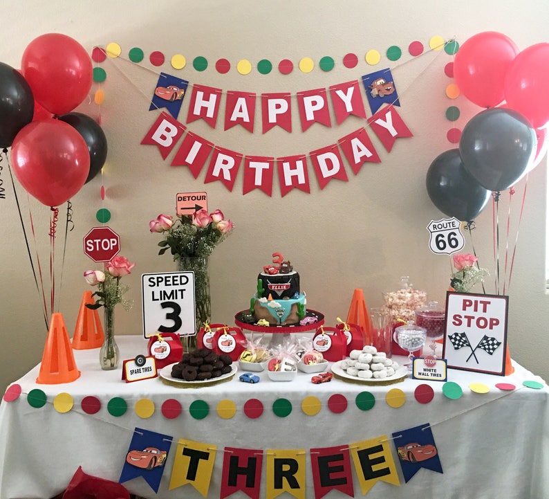 May include: A Cars-themed birthday party table setting with red and black balloons, a cake with a race car on top, and a banner that says "Happy Birthday". There are also various decorations, including traffic signs, cones, and a banner that says "Three".