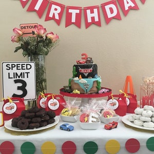 May include: A Cars-themed birthday party table setting with a two-tier cake decorated with a tire, a cactus, and the characters Lightning McQueen and Mater. The cake is on a white cake stand. There are also donuts, cookies, and other treats on the table. The table is decorated with red, yellow, and green circles.