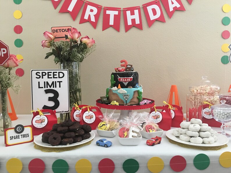 May include: A Cars-themed birthday party table setting with a two-tier cake decorated with a tire, a cactus, and the characters Lightning McQueen and Mater. The cake is on a white cake stand. There are also donuts, cookies, and other treats on the table. The table is decorated with red, yellow, and green circles.