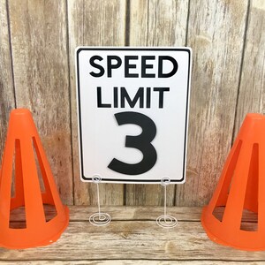 May include: A white speed limit sign with black text that reads "SPEED LIMIT 3". The sign is propped up on a white metal stand. Two orange traffic cones are on either side of the sign.