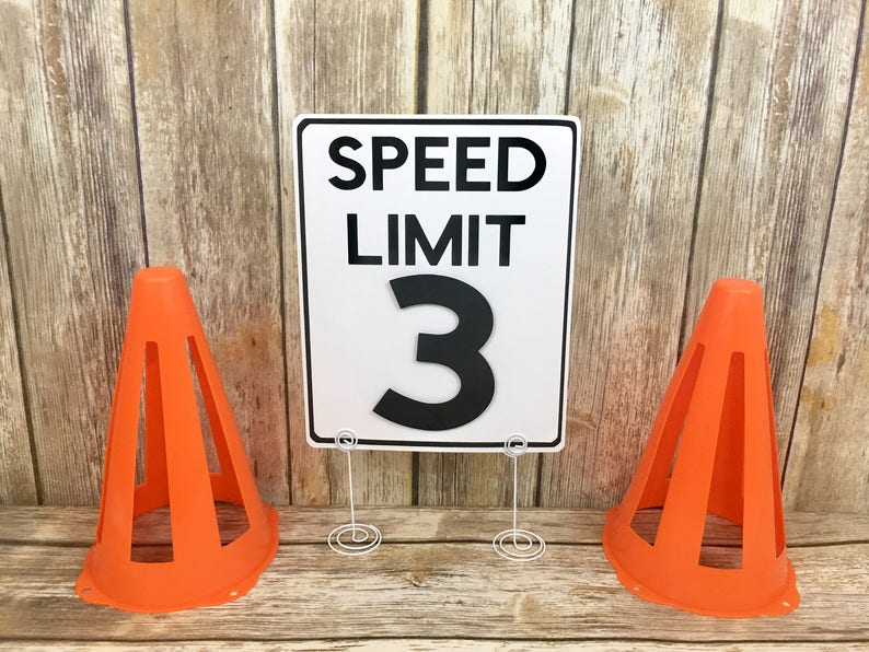 May include: A white speed limit sign with black text that reads "SPEED LIMIT 3". The sign is propped up on a white metal stand. Two orange traffic cones are on either side of the sign.