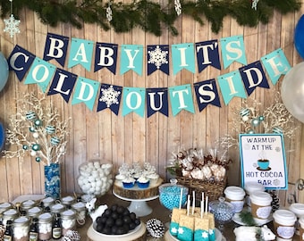 May include: A winter-themed baby shower table with blue and white decorations. The table is decorated with a banner that says "Baby It's Cold Outside". There are also cupcakes, candy, and hot cocoa bar signs.