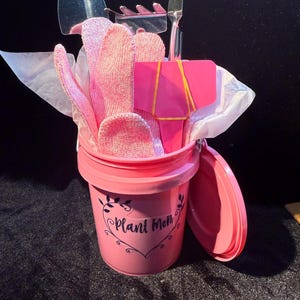 May include: A pink gardening gift set in a small bucket. The bucket has the words "Plant Mom" in a heart-shaped design. The set includes gardening tools, pink gloves, and a pink plant marker.