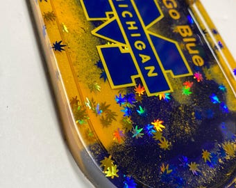 May include: An oval resin tray featuring a deep blue and yellow color scheme with embedded holographic leaf-shaped glitter. The center displays the block M logo with the text MICHIGAN and the phrase Go Blue in yellow lettering on a blue background.