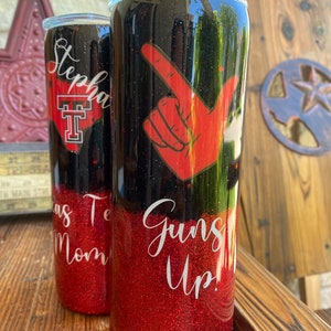 May include: Two red and black glitter tumblers with a white design. One tumbler has the text "Guns Up!" and the other has the text "Texas Mom".