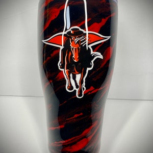May include: A black and red stainless steel tumbler with a white and orange logo of a horse and rider. The logo is a silhouette of a rider wearing a hat and cape, riding a horse. The tumbler has a black lid with a clear plastic lid.