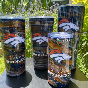May include: Four Denver Broncos themed tumblers with a black, white, orange, and brown marbled design. The tumblers have glitter and the Denver Broncos logo.
