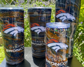 May include: Four Denver Broncos themed tumblers with a black, white, orange, and brown marbled design. The tumblers have glitter and the Denver Broncos logo.