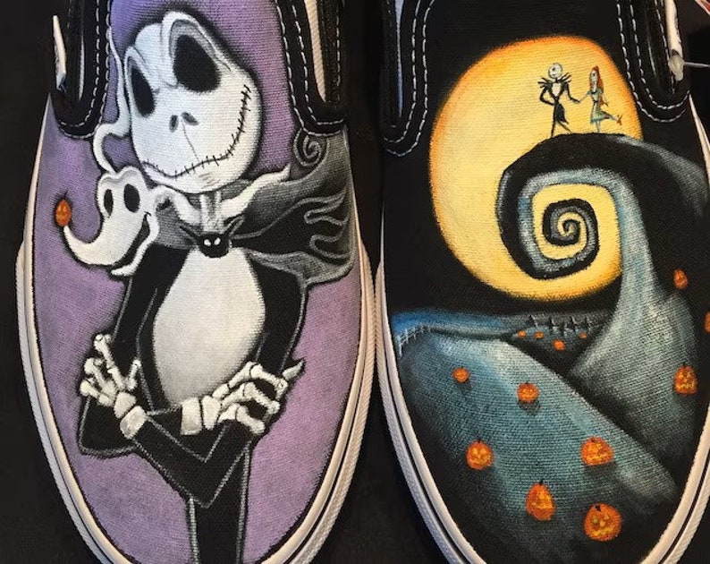 Op de afbeelding: Een paar op maat geverfde Vans slip-on schoenen met personages uit de film The Nightmare Before Christmas. De ene schoen heeft Jack Skellington, de Pumpkin King, op een paarse achtergrond. De andere schoen heeft Jack en Sally op een heuvel met een volle maan en een spiraalvormige heuvel op de achtergrond.