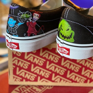 Op de afbeelding: Een paar zwarte Vans sneakers met een aangepast handgeschilderd ontwerp met personages uit The Nightmare Before Christmas. De linker schoen heeft Jack Skellington, Sally en Oogie Boogie. De rechter schoen heeft een close-up van Jack Skellington's gezicht.
