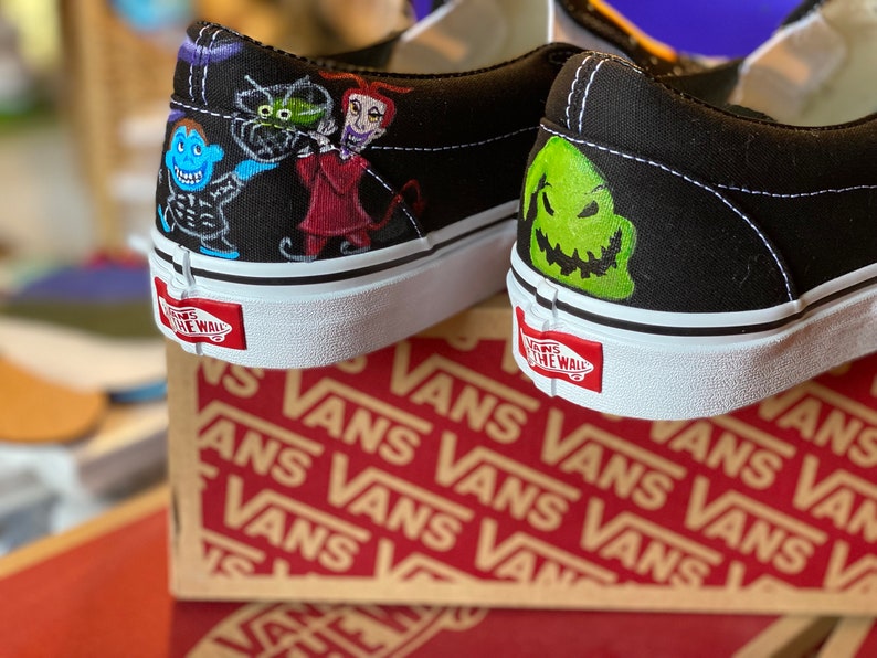 Op de afbeelding: Een paar zwarte Vans sneakers met een aangepast handgeschilderd ontwerp met personages uit The Nightmare Before Christmas. De linker schoen heeft Jack Skellington, Sally en Oogie Boogie. De rechter schoen heeft een close-up van Jack Skellington's gezicht.