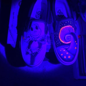 Op de afbeelding: Een paar zwarte Vans slip-ons met glow-in-the-dark verf. De ene schoen heeft een Jack Skellington ontwerp, terwijl de andere een spiraalvormig ontwerp heeft met sterren en pompoenen.