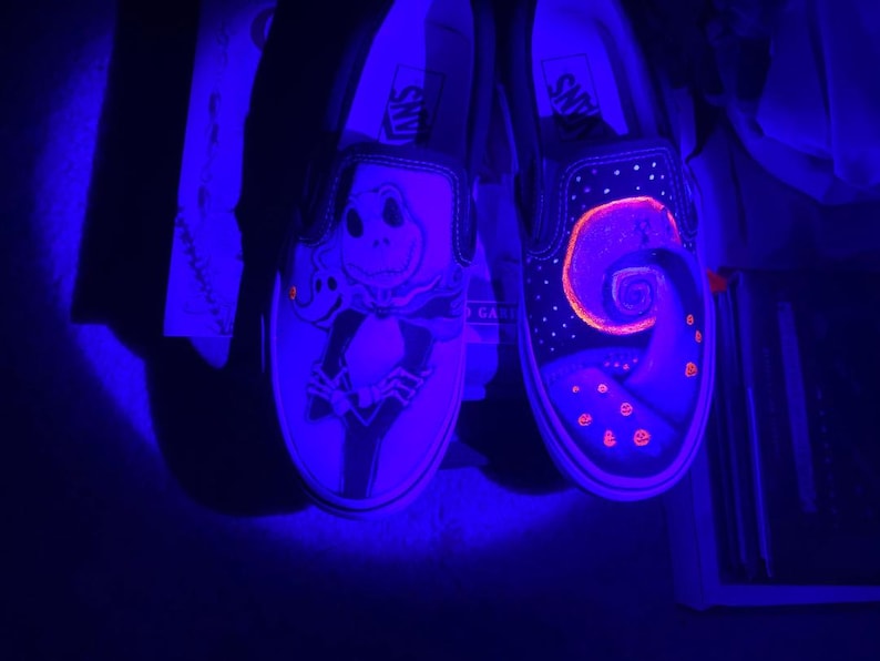 Op de afbeelding: Een paar zwarte Vans slip-ons met glow-in-the-dark verf. De ene schoen heeft een Jack Skellington ontwerp, terwijl de andere een spiraalvormig ontwerp heeft met sterren en pompoenen.