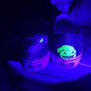 Op de afbeelding: Een paar zwarte canvas schoenen met glow-in-the-dark ontwerpen. De ene schoen heeft een groen en geel Jack Skellington ontwerp, terwijl de andere schoen een groen en geel Oogie Boogie ontwerp heeft.