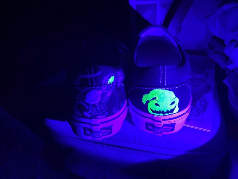 Op de afbeelding: Een paar zwarte canvas schoenen met glow-in-the-dark ontwerpen. De ene schoen heeft een groen en geel Jack Skellington ontwerp, terwijl de andere schoen een groen en geel Oogie Boogie ontwerp heeft.