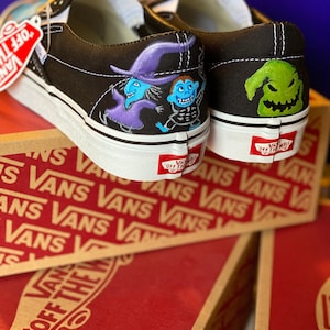 Op de afbeelding: Een paar zwarte Vans slip-on schoenen met aangepaste geschilderde ontwerpen. De ene schoen heeft een paarse en blauwe heks en een blauw skeletpersonage. De andere schoen heeft een groen, grijnzend monster met een puntige hoed. De schoenen staan op een doos met het Vans-logo.