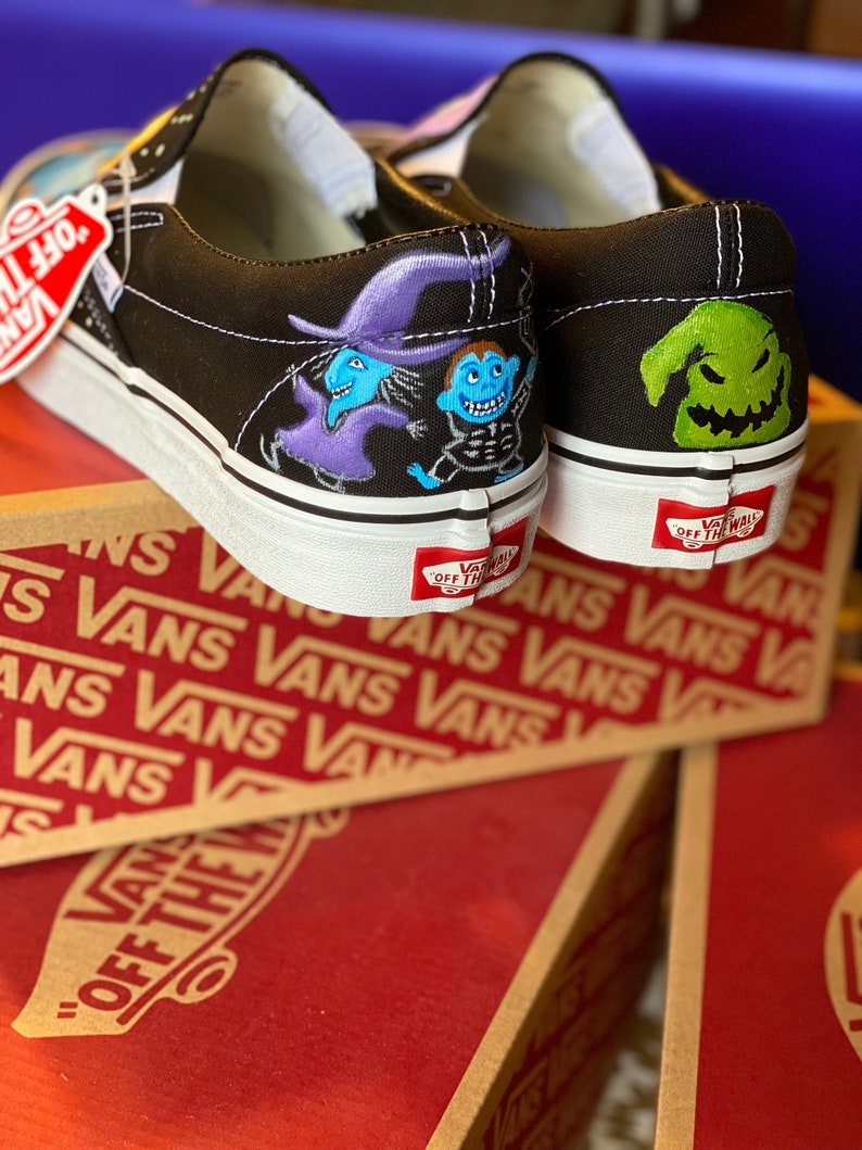 Op de afbeelding: Een paar zwarte Vans slip-on schoenen met aangepaste geschilderde ontwerpen. De ene schoen heeft een paarse en blauwe heks en een blauw skeletpersonage. De andere schoen heeft een groen, grijnzend monster met een puntige hoed. De schoenen staan op een doos met het Vans-logo.