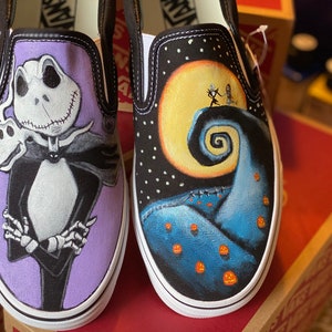 Op de afbeelding: Een paar op maat geschilderde Vans slip-on schoenen met personages uit de film The Nightmare Before Christmas. De ene schoen heeft Jack Skellington op een paarse achtergrond, terwijl de andere schoen het iconische Halloween Town landschap met Jack en Sally heeft.