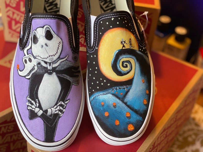 Op de afbeelding: Een paar op maat geschilderde Vans slip-on schoenen met personages uit de film The Nightmare Before Christmas. De ene schoen heeft Jack Skellington op een paarse achtergrond, terwijl de andere schoen het iconische Halloween Town landschap met Jack en Sally heeft.