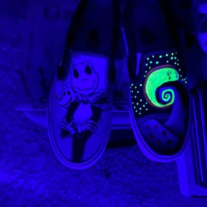 Op de afbeelding: Een paar zwarte slip-on schoenen met glow-in-the-dark verf. De ene schoen heeft een groen en zwart Jack Skellington ontwerp, terwijl de andere schoen een groen en zwart Nightmare Before Christmas ontwerp heeft.