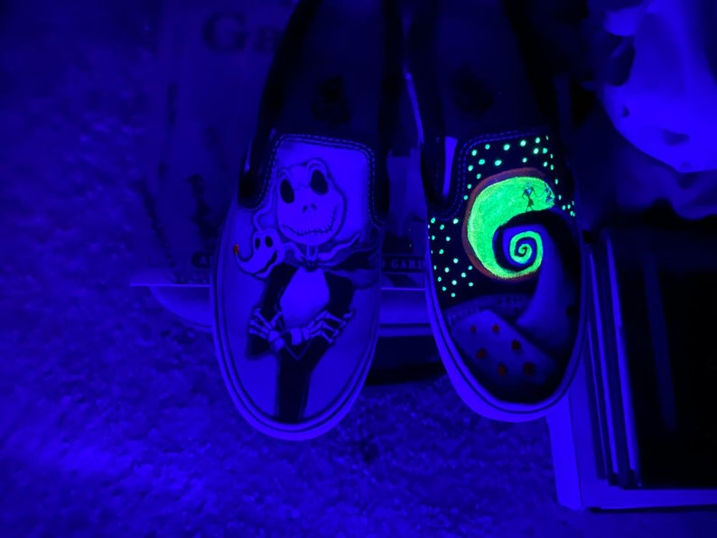 Op de afbeelding: Een paar zwarte slip-on schoenen met glow-in-the-dark verf. De ene schoen heeft een groen en zwart Jack Skellington ontwerp, terwijl de andere schoen een groen en zwart Nightmare Before Christmas ontwerp heeft.