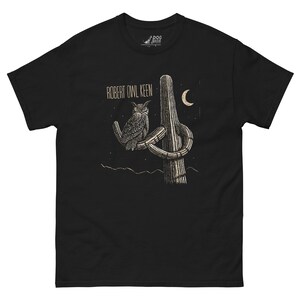 May include: Black t-shirt with a graphic of an owl perched on a cactus with a crescent moon in the background. The text "ROBERT OWL KEEN" is printed on the shirt.