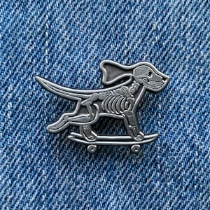 May include: A silver-toned enamel pin of a skeleton dog skateboarding. The dog is in a running pose with its tongue sticking out. The skateboard has wheels and is on a denim background.