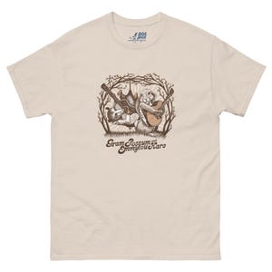 Könnte beinhalten: Hellbeige T-Shirt mit einer Grafik eines Opossums und eines Hasen, die Gitarre spielen, umgeben von einem Astdesign. Der Text "Gram Possum and Emmylou Hare" ist darunter gedruckt. Das T-Shirt hat einen klassischen Rundhalsausschnitt.