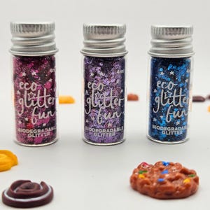 May include: Three small glass bottles filled with colourful glitter. Each bottle has a silver cap and features the text "eco glitter fun" and "biodegradable glitter". The glitter colours are pink, purple, and blue.