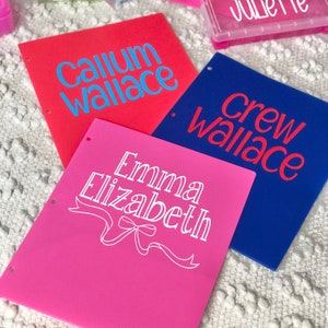 May include: Three colorful binders: red with "Callum Wallace" in blue, blue with "Crew Wallace" in red, and pink with "Emma Elizabeth" in white. Each binder has three holes for insertion into a binder.