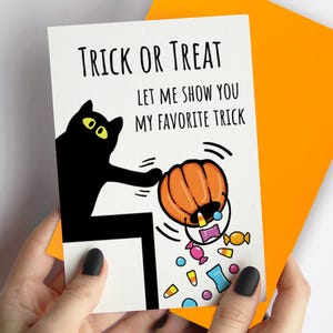 May include: A white greeting card featuring a black cat illustration and the text Trick or Treat, Let me show you my favourite trick. The cat is tipping over an orange pumpkin bucket, spilling colourful sweets onto the bottom of the card.