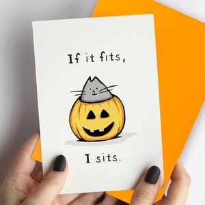 May include: A white greeting card featuring a hand-drawn illustration of a gray cat sitting inside a carved orange jack-o'-lantern pumpkin. The text "If it fits, I sits." is printed in black ink above and below the cat. A bright orange envelope is behind the card.