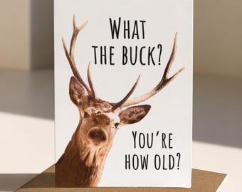 Puede incluir: Una tarjeta de felicitación blanca con un ciervo marrón con astas y el texto negro WHAT THE BUCK? YOU'RE HOW OLD? La tarjeta está colocada en posición vertical sobre un sobre marrón, mostrando un diseño claro y contrastado centrado en el animal.