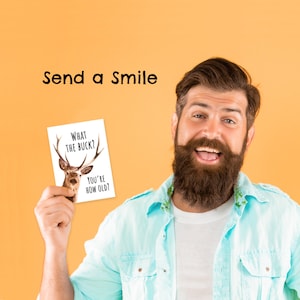 May include: A man with a beard is holding a card that says "What the buck? You're how old?" The card has a picture of a deer on it. The man is smiling and looking at the camera. The background is a solid orange color. Send a smile.