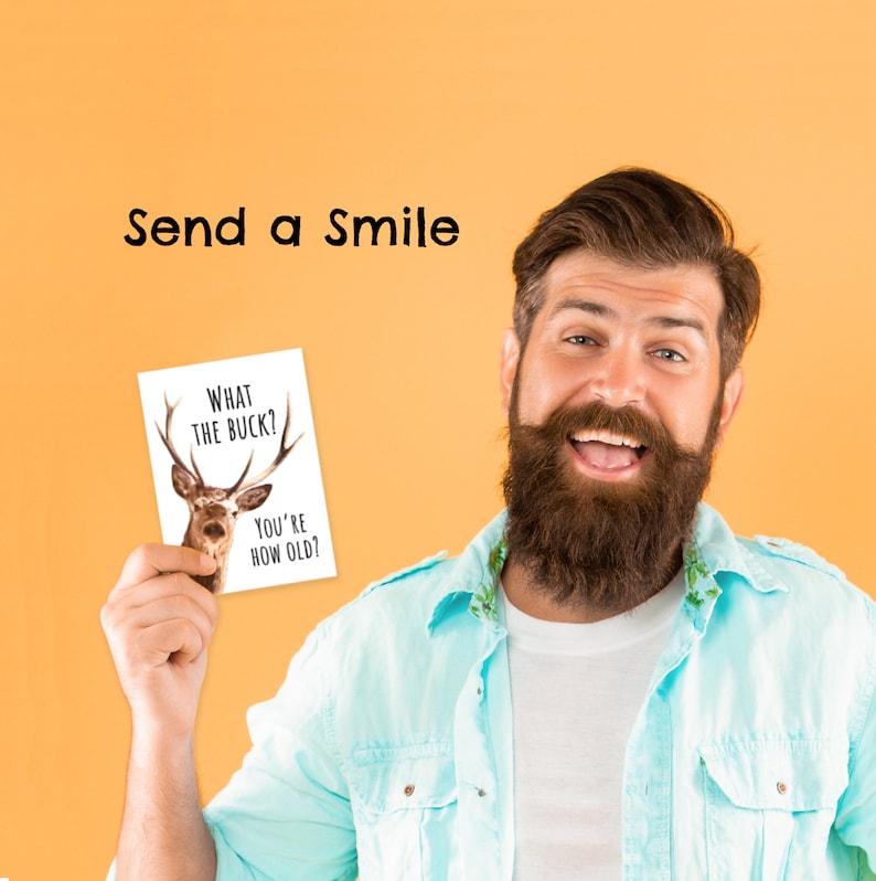 May include: A man with a beard is holding a card that says "What the buck? You're how old?" The card has a picture of a deer on it. The man is smiling and looking at the camera. The background is a solid orange color. Send a smile.