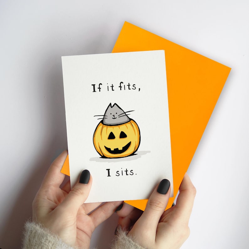 May include: A white greeting card featuring a hand-drawn illustration of a gray cat sitting inside a carved orange jack-o'-lantern pumpkin. The text "If it fits, I sits." is printed in black ink above and below the cat. A bright orange envelope is behind the card.