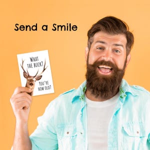 May include: A white rectangular greeting card featuring a graphic of a deer with large antlers and the black text "WHAT THE BUCK? YOU'RE HOW OLD?" held up against a solid orange background with the black text "Send a Smile" above.