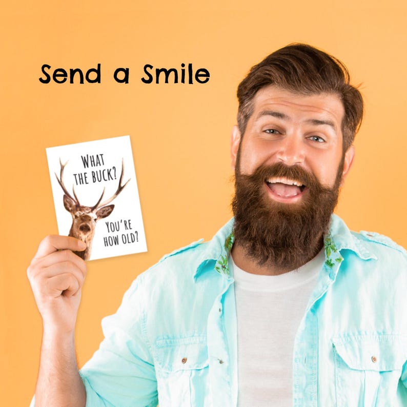 May include: A white rectangular greeting card featuring a graphic of a deer with large antlers and the black text "WHAT THE BUCK? YOU'RE HOW OLD?" held up against a solid orange background with the black text "Send a Smile" above.