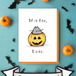 May include: A white greeting card featuring a cartoon illustration of a gray cat sitting inside a carved orange jack-o'-lantern pumpkin. The text reads "If it fits, I sits." The card is layered over an orange envelope, with a banner below stating "Spooky season is here!"