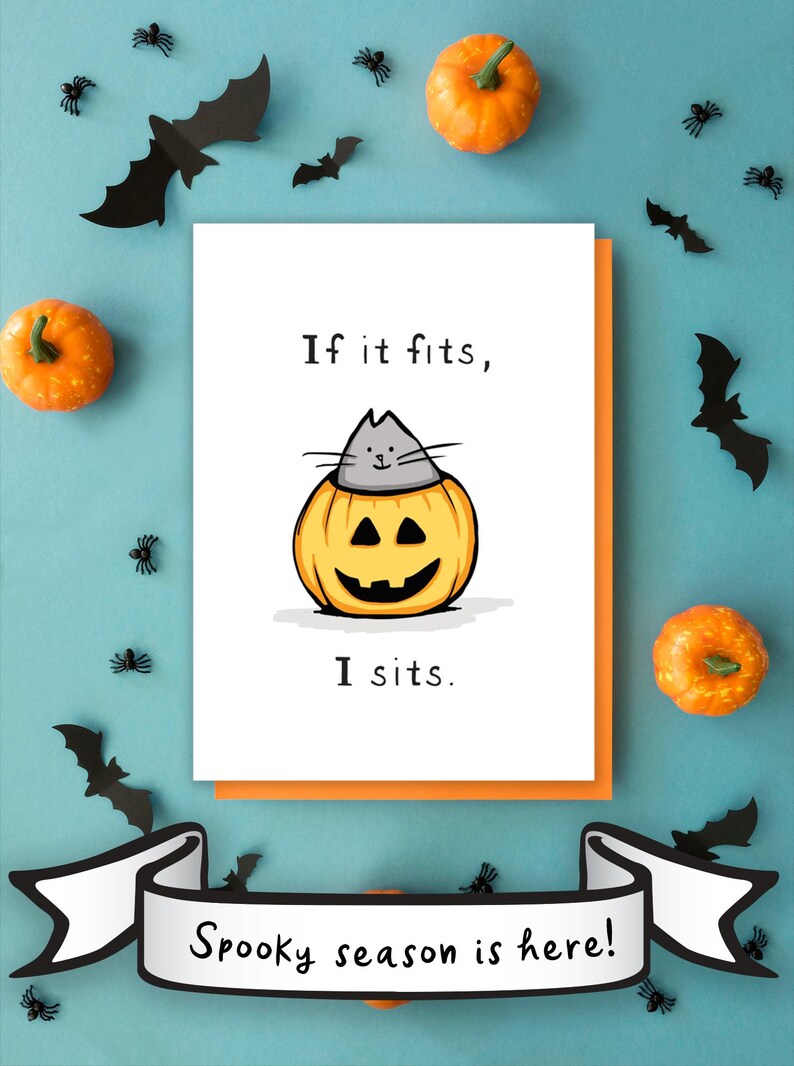May include: A white greeting card featuring a cartoon illustration of a gray cat sitting inside a carved orange jack-o'-lantern pumpkin. The text reads "If it fits, I sits." The card is layered over an orange envelope, with a banner below stating "Spooky season is here!"