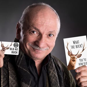 May include: A rectangular card featuring a deer illustration and the text "OLD AS BUCK" and a vertical card featuring a deer illustration and the text "WHAT THE BUCK? YOU'RE HOW OLD?" printed in black ink on a white background.