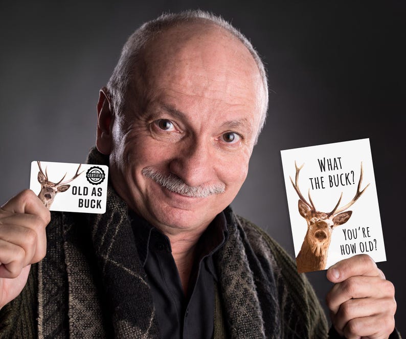 May include: A rectangular card featuring a deer illustration and the text "OLD AS BUCK" and a vertical card featuring a deer illustration and the text "WHAT THE BUCK? YOU'RE HOW OLD?" printed in black ink on a white background.