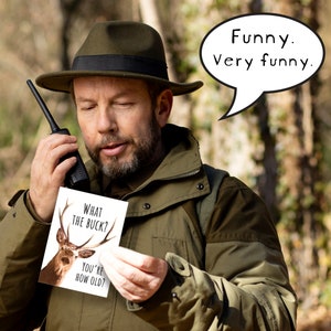 May include: A man in a green jacket and a brown hat holds a card with a deer on it. The card says "What the buck? You're how old?" A speech bubble above him says "Funny. Very funny."