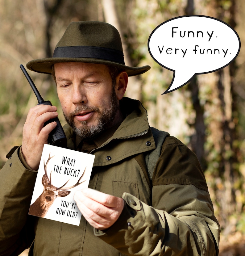 May include: A man in a green jacket and a brown hat holds a card with a deer on it. The card says "What the buck? You're how old?" A speech bubble above him says "Funny. Very funny."