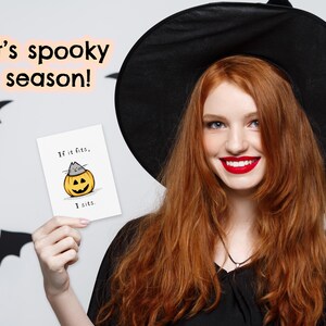 May include: A white greeting card featuring a cartoon illustration of a gray cat sitting inside a carved orange jack-o'-lantern pumpkin. The text on the card reads, "If it fits, I sits." The card is held up against a background with bat silhouettes.