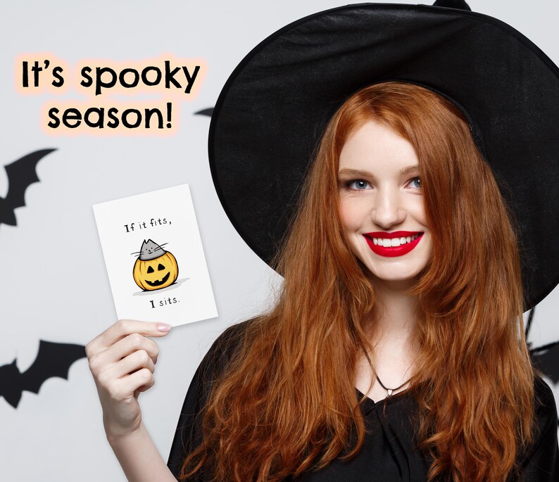 May include: A white greeting card featuring a cartoon illustration of a gray cat sitting inside a carved orange jack-o'-lantern pumpkin. The text on the card reads, "If it fits, I sits." The card is held up against a background with bat silhouettes.