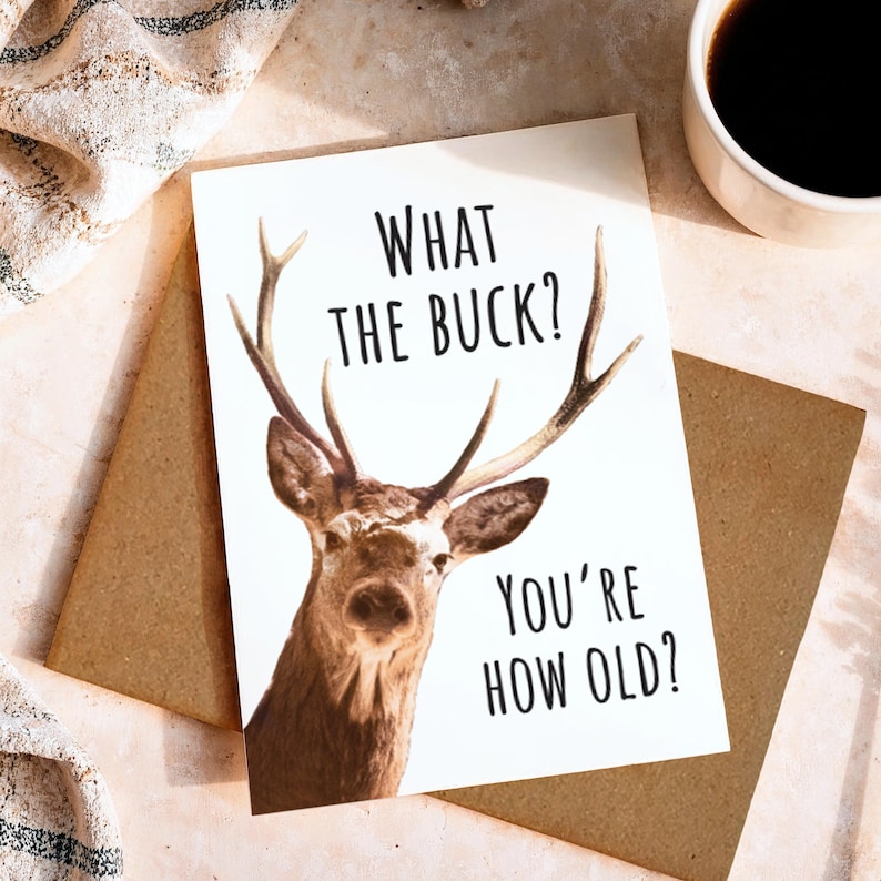 May include: A white greeting card featuring a brown deer with antlers and the black text "WHAT THE BUCK? YOU'RE HOW OLD?" printed on the front. The card is positioned on top of a brown envelope, with a white mug of coffee visible in the corner.