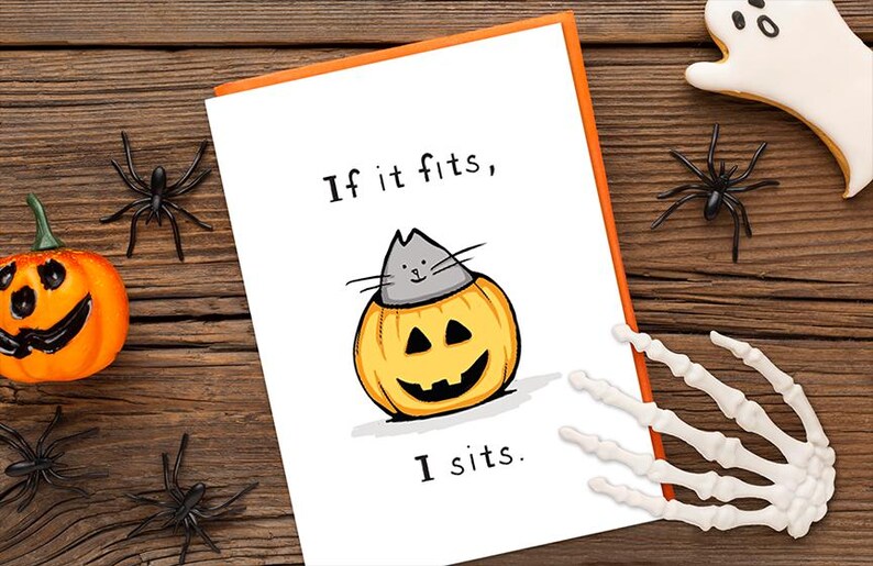 May include: A white greeting card with an orange border featuring a cartoon illustration of a gray cat sitting inside a carved orange jack-o'-lantern pumpkin. The text on the card reads, If it fits, I sits. The card is surrounded by Halloween decor.