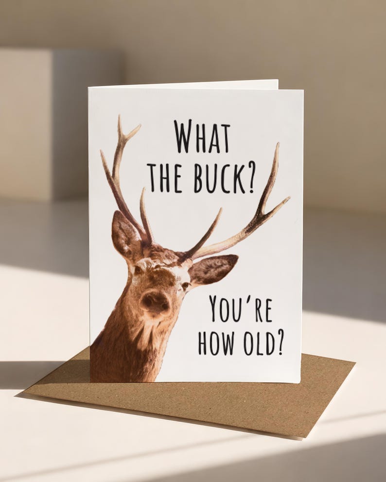 May include: A white greeting card featuring a brown deer with antlers and the black text WHAT THE BUCK? YOU'RE HOW OLD? The card is positioned upright on a brown envelope, displaying a clear, high-contrast design with a focus on the animal graphic.